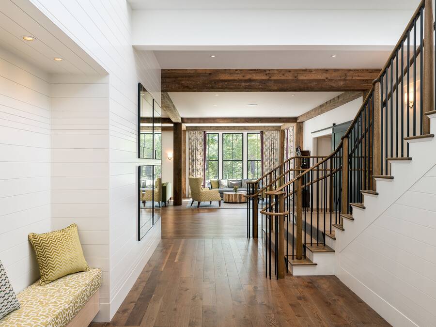 A spacious interior with a wooden staircase, a seating area with patterned cushions, and large windows allowing natural light.