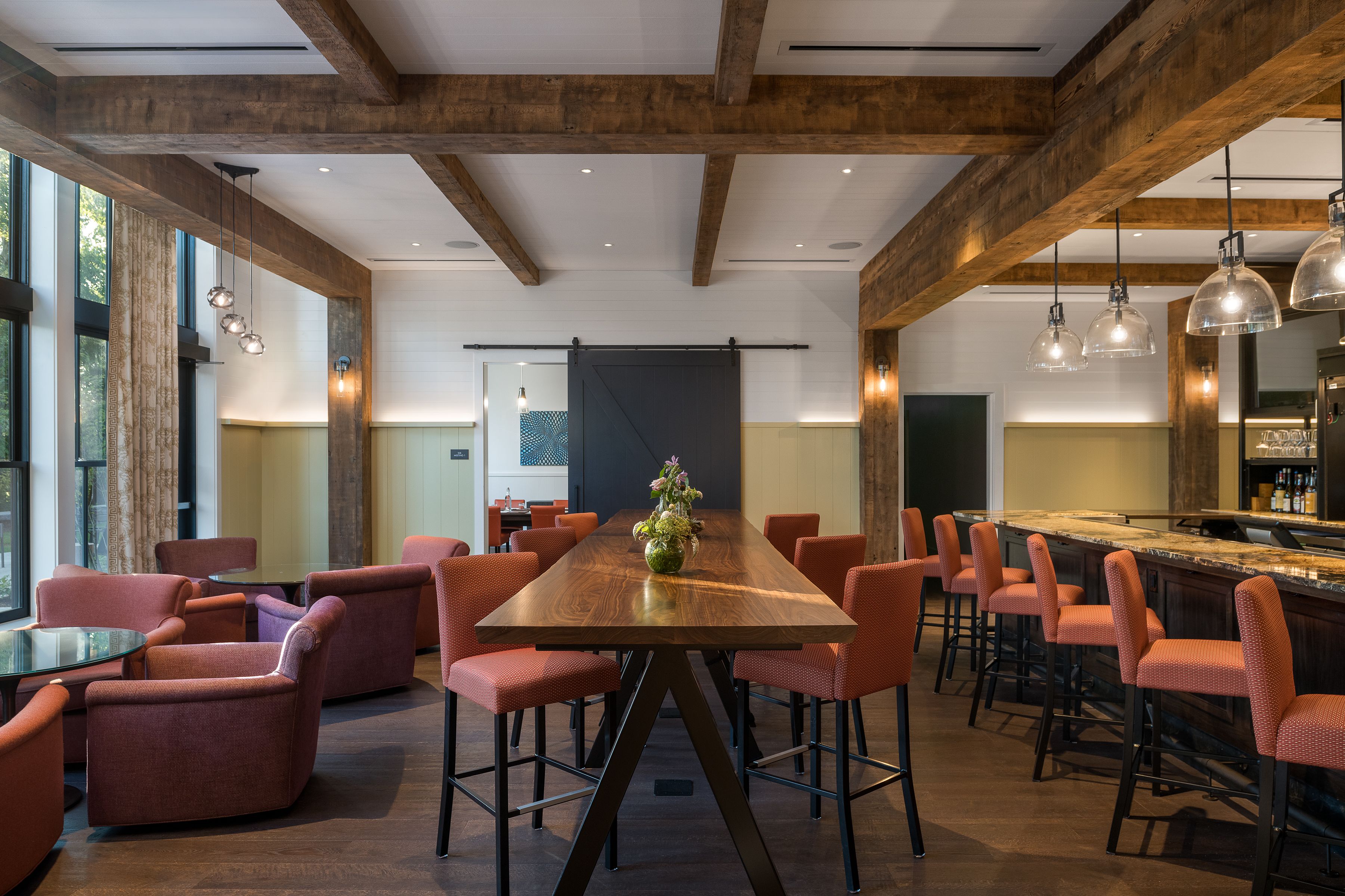 A spacious dining area with a long wooden table surrounded by red upholstered chairs, a bar with stools, and pendant lighting.