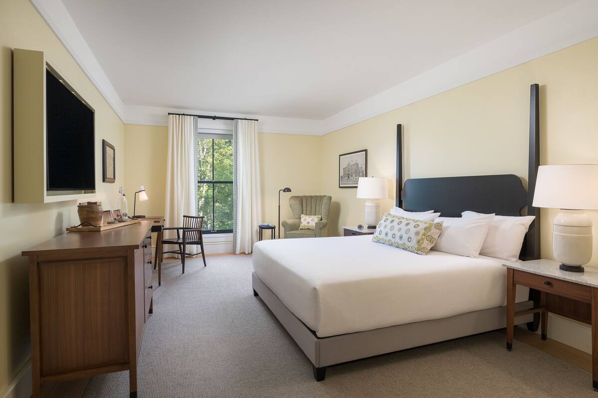 A hotel room with a large bed, a desk, a chair, a window with curtains, and a nightstand with a lamp.
