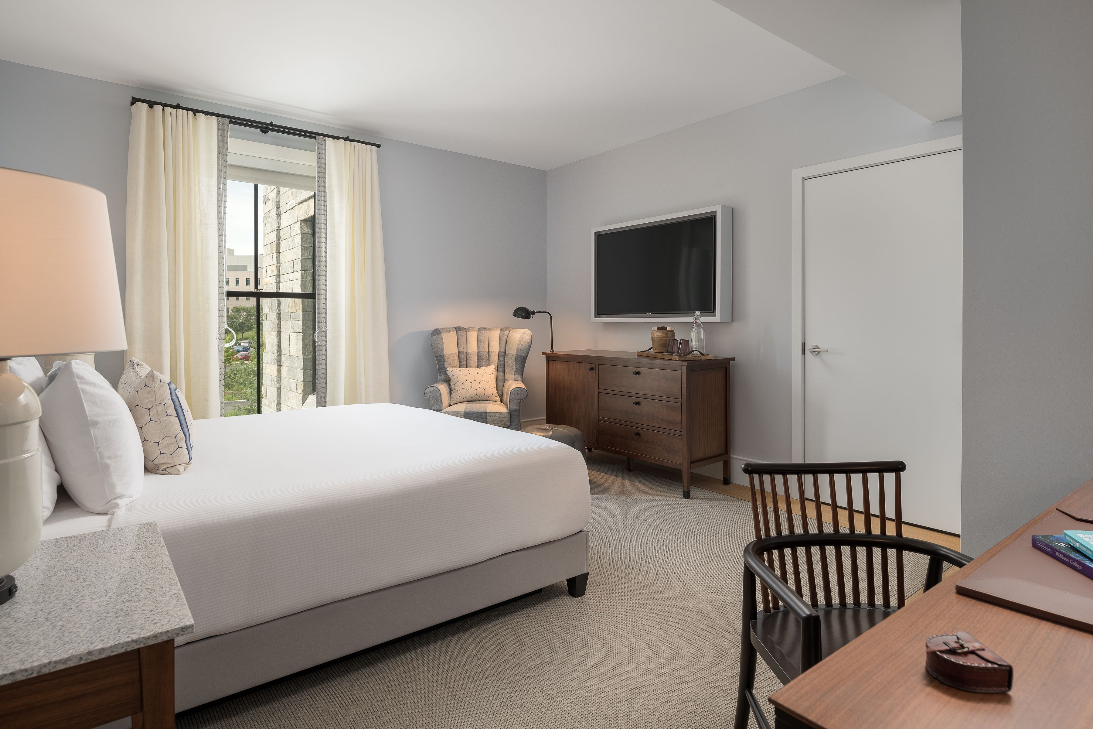 A hotel room with a bed, a desk, a chair, a TV, and a window with curtains.