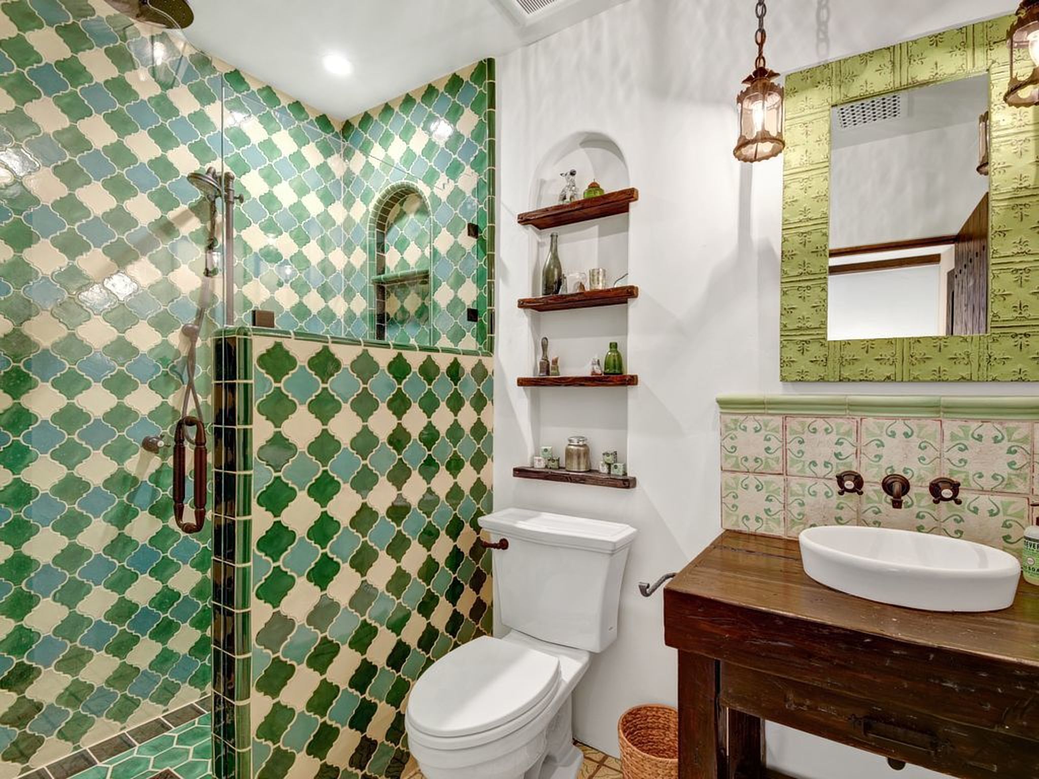 Un baño con un patrón de baldosas blancas y verdes a cuadros, un inodoro blanco, un tocador de madera con un lavamanos blanco y una lámpara colgante.