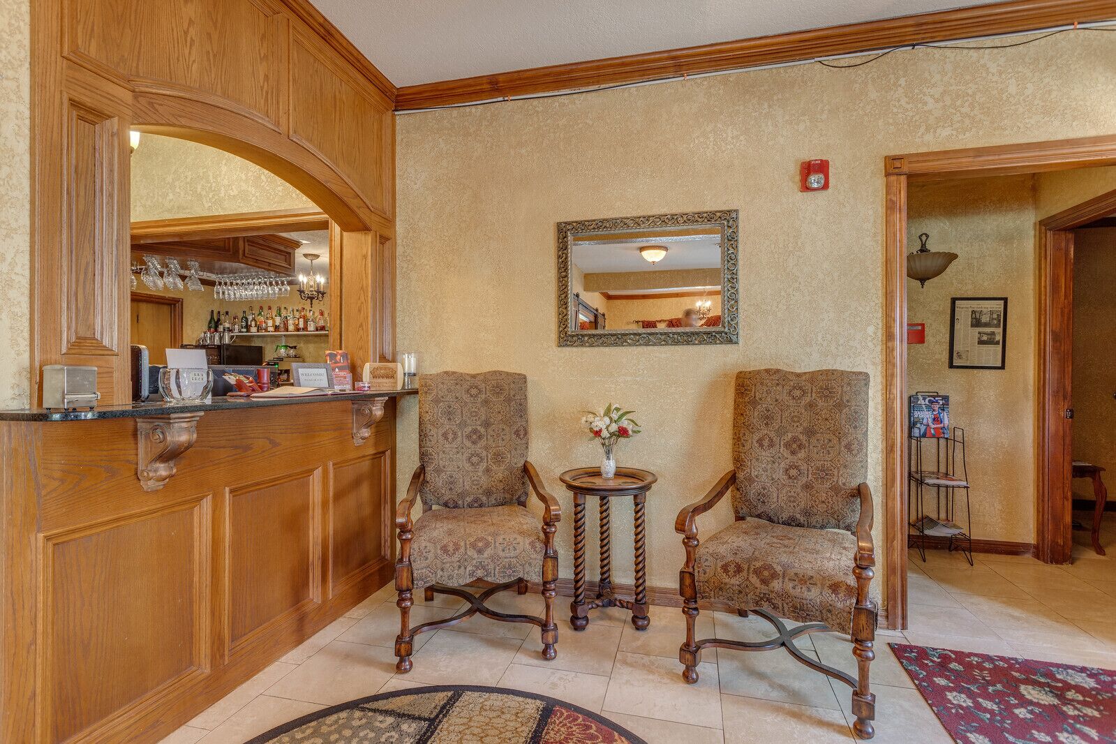 Un hall d’entrée avec des boiseries, deux fauteuils à motifs, une petite table ronde surmontée d’un vase, un miroir au mur et, au fond, un coin bar.