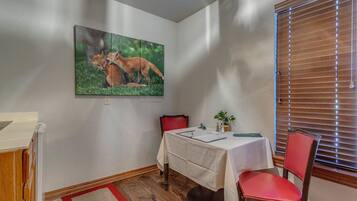 A dining area with a table set for two, a painting of foxes on the wall, and a window with blinds.