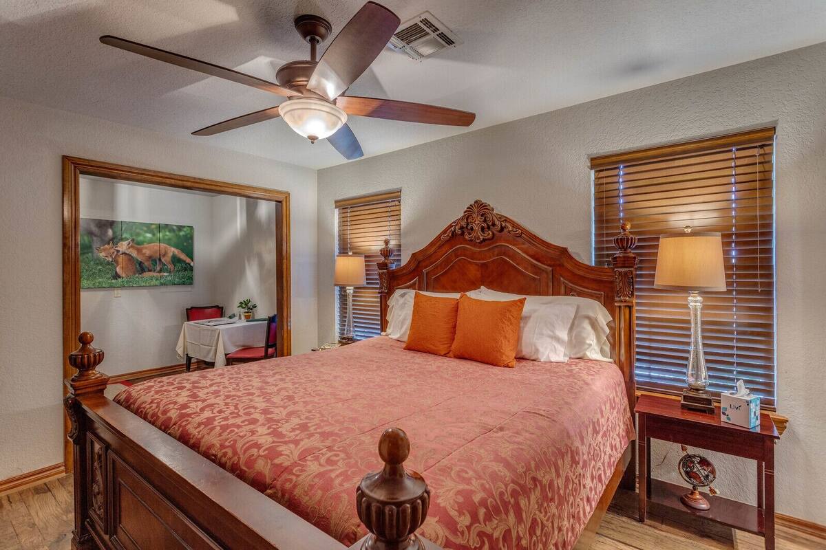 A hotel room with a wooden bed, a ceiling fan, a desk with a chair, a table with a lamp, and a painting on the wall.
