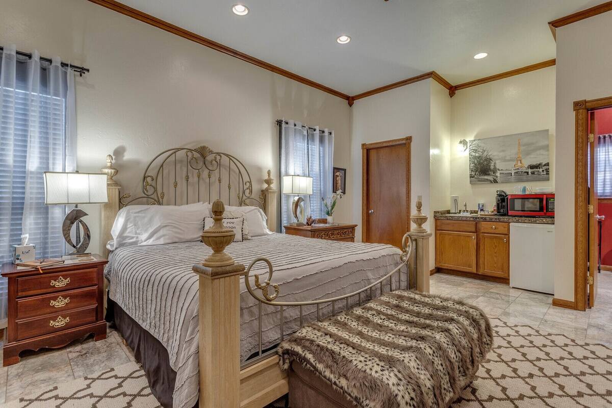 A bedroom with a large bed, bedside tables, a nightstand, a wooden wardrobe, a bathroom with a shower, and a kitchen area with a microwave.