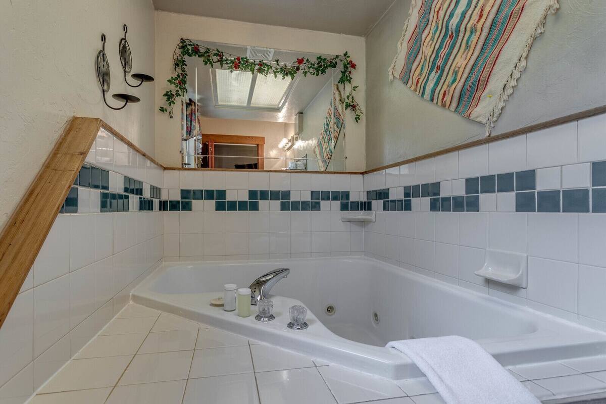 A bathroom with a large bathtub, white tiled walls, a mirror, and a wooden frame on the left.
