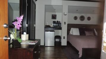 A compact hotel room with a bed, a small kitchenette, a trash bin, and a wooden dresser with a vase of flowers.