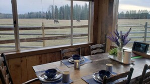 A wooden dining table with blue placemats and bowls, set against a window overlooking a fenced outdoor area with horses. - Woodhouse Cottages and Ranch (Prince George)