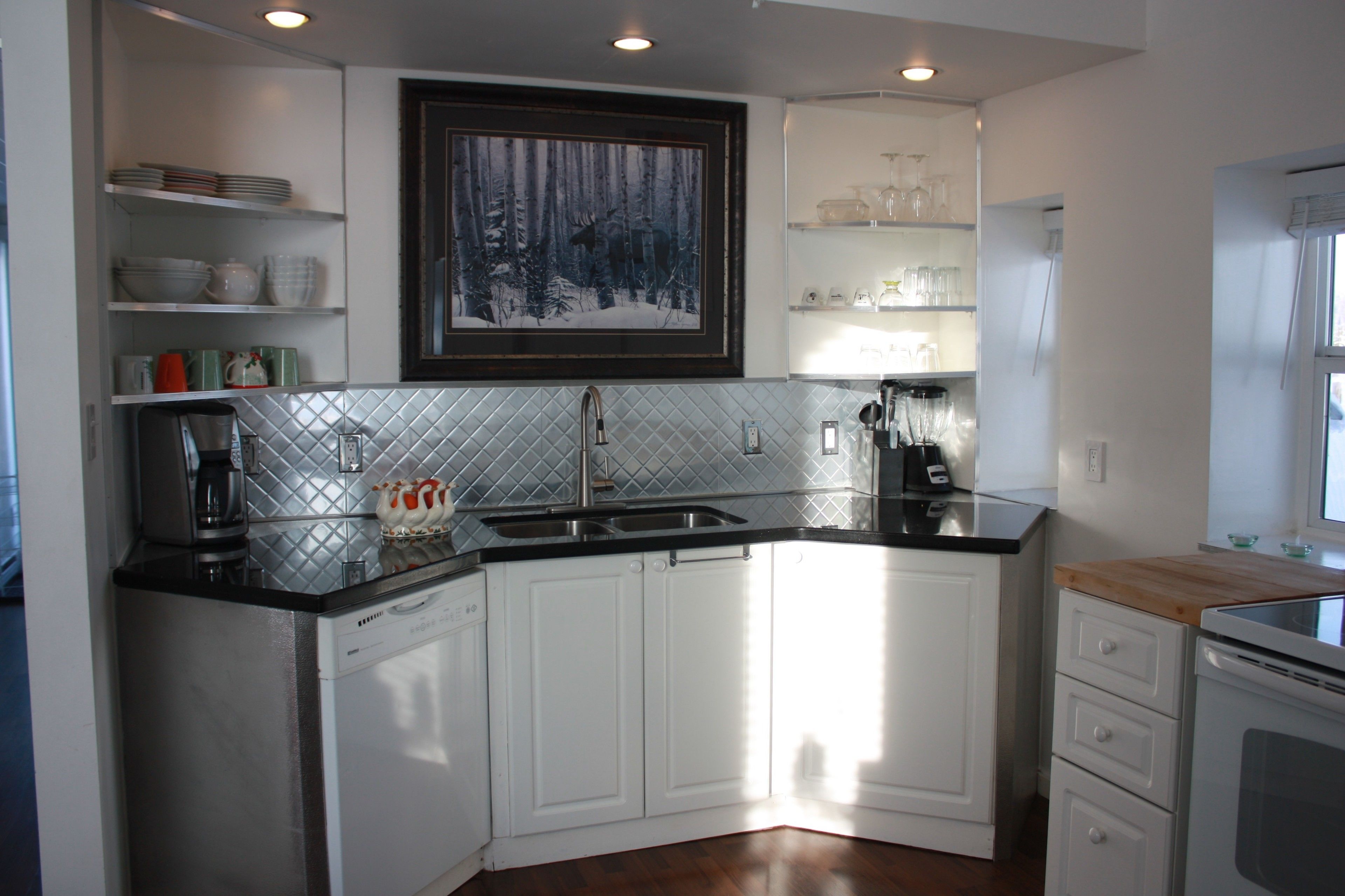 A modern kitchen with white cabinetry, a black countertop, and a diamond-patterned backsplash. There is a painting of a forest scene above the sink and shelves with dishes and glasses.