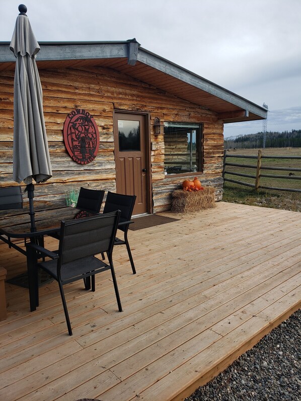 Una cabaña rústica con una terraza de madera, una mesa de comedor con sillas y una puerta con ventanal.
