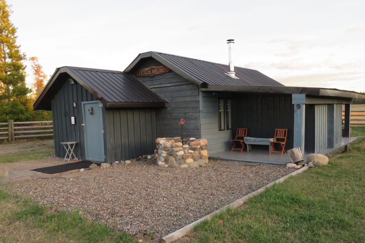 A rustic building with a stone foundation, a metal roof, and a sign reading "Little Muddy." There are two wooden chairs outside.