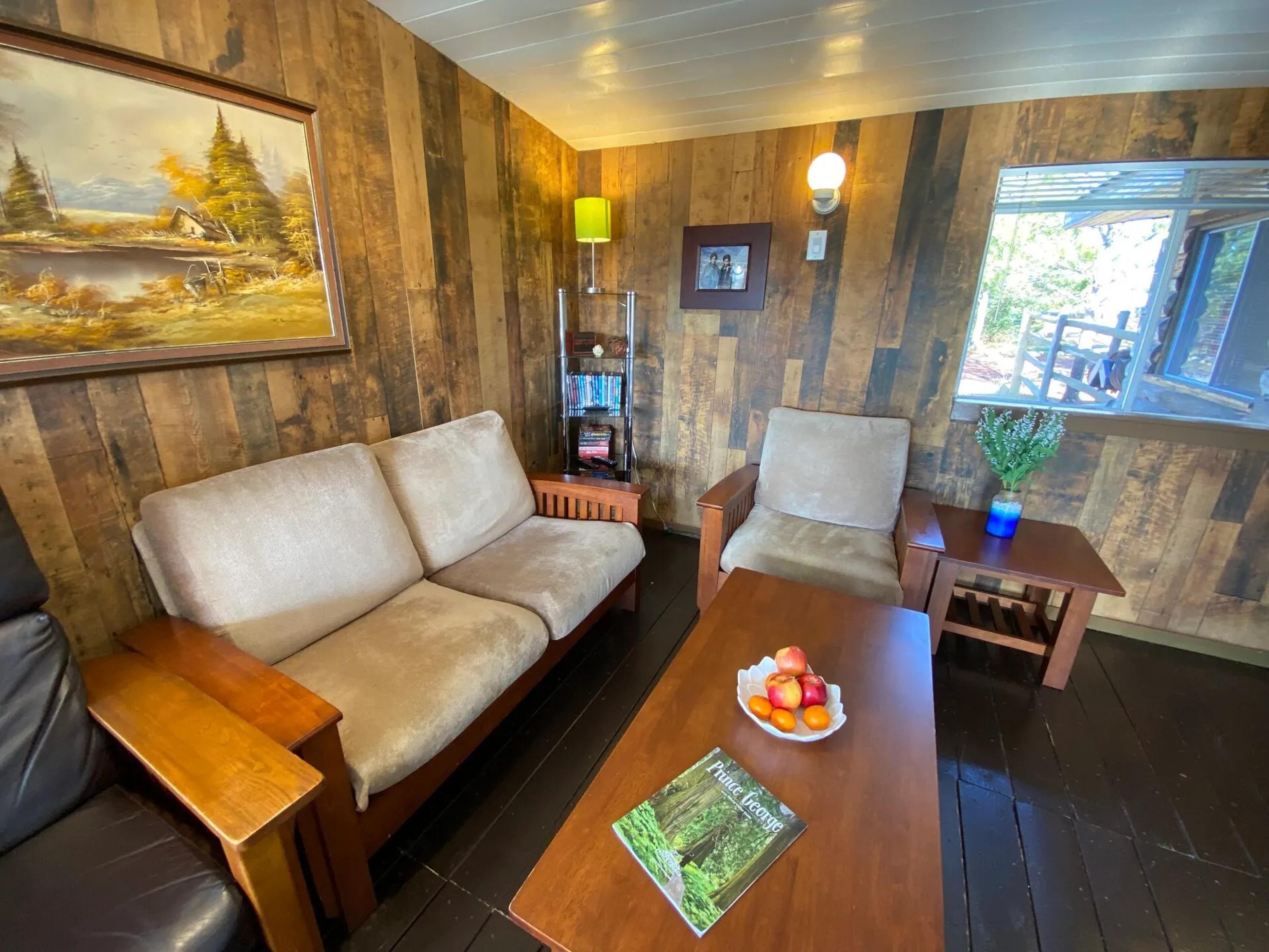 A cozy room with a wooden wall, a sofa, a chair, a small table with a book and fruit, and a bookshelf.