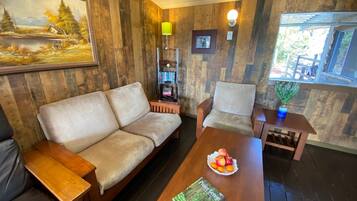 A cozy room with a wooden wall, a sofa, a chair, a small table with a book and fruit, and a bookshelf.