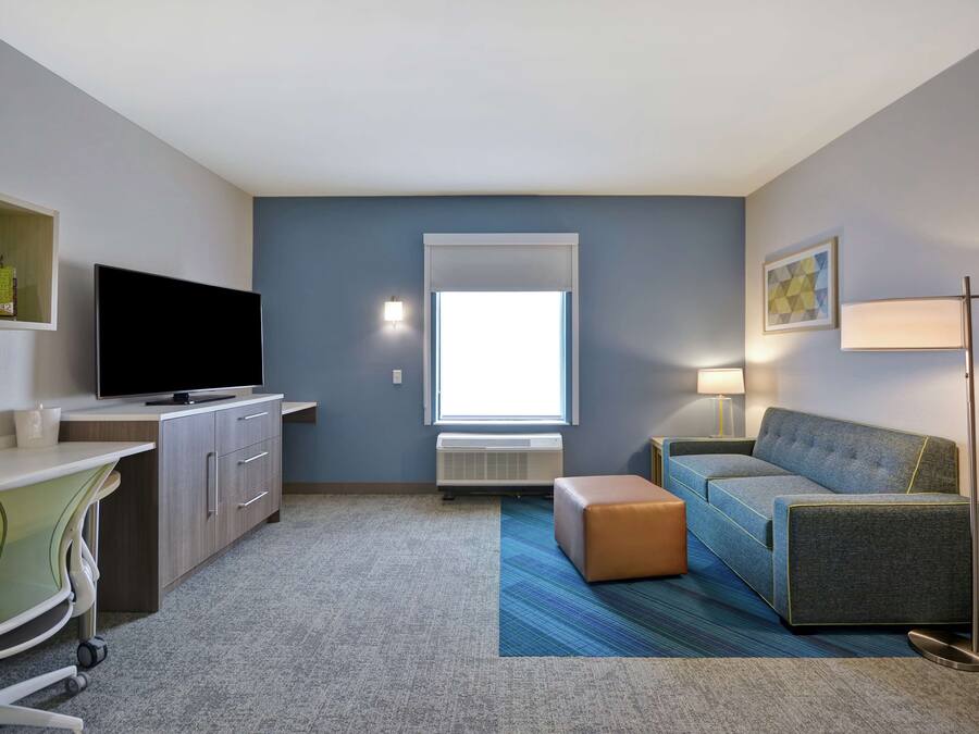 Habitación de hotel compacta con televisor de pantalla plana, escritorio con silla, sofá, lámpara y ventana con persianas.