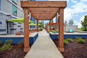 A covered outdoor seating area with tables and chairs, a concrete pathway, and a building with a stone facade. - Home2 Suites by Hilton Atlanta Marietta (Marietta)