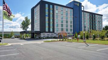 Un hotel moderno con un llamativo letrero de "HOME 2 SUITES BY HILTON", un amplio estacionamiento y un asta con la bandera estadounidense.