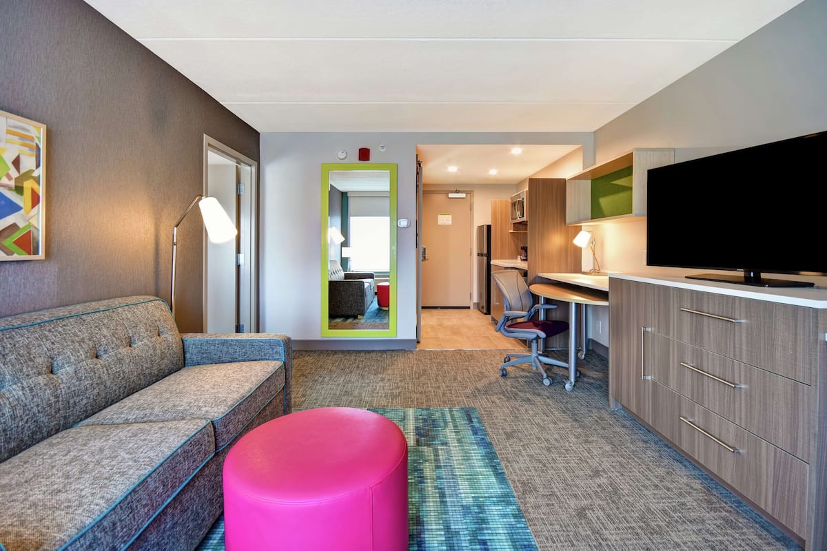 Une chambre d’hôtel moderne équipée d’un canapé, d’un pouf rose, d’un téléviseur à écran plat, d’un bureau et d’un miroir.