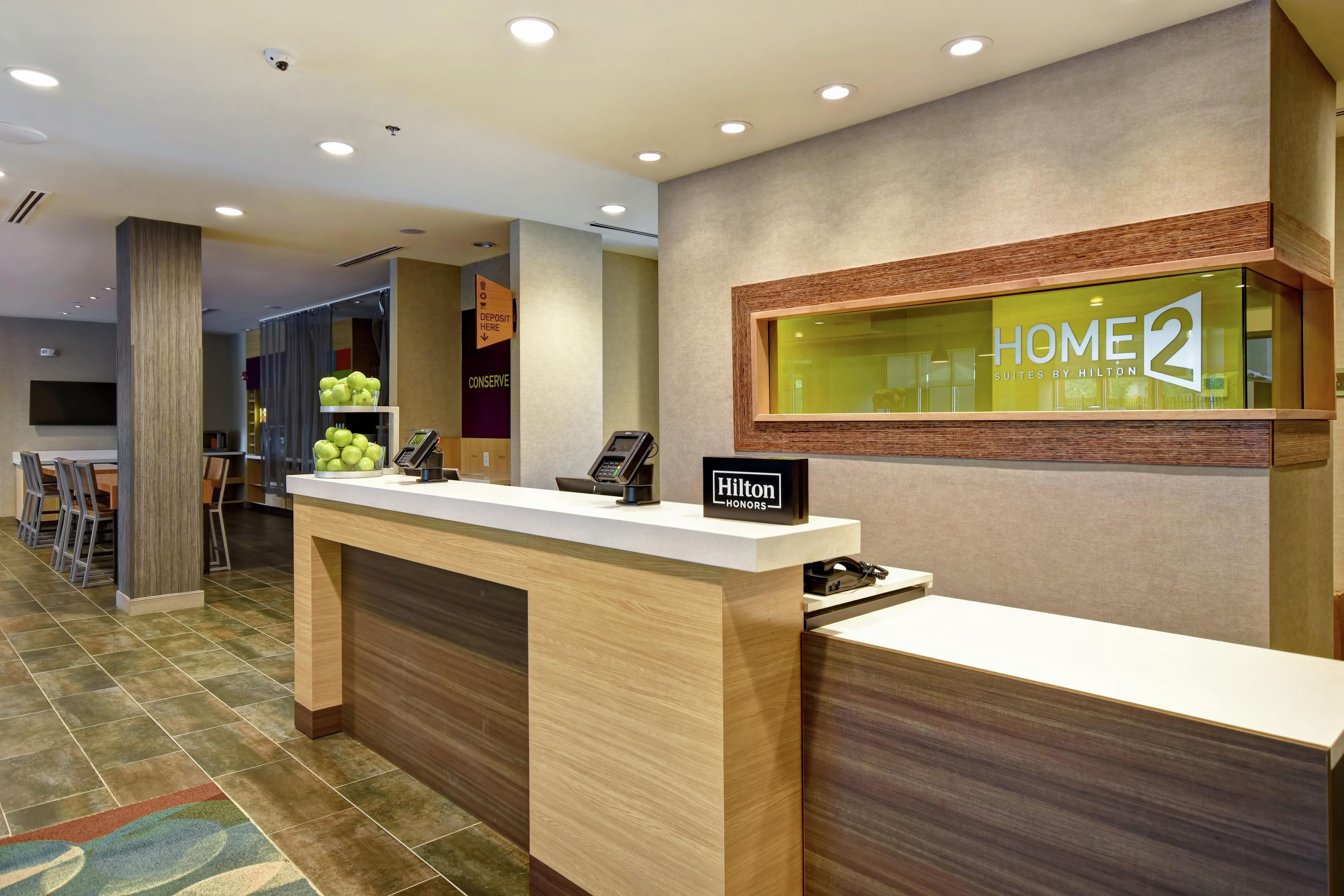 A hotel reception desk with a 'Home2' sign, a 'Hilton Honors' sign, and a 'Conserve' sign.