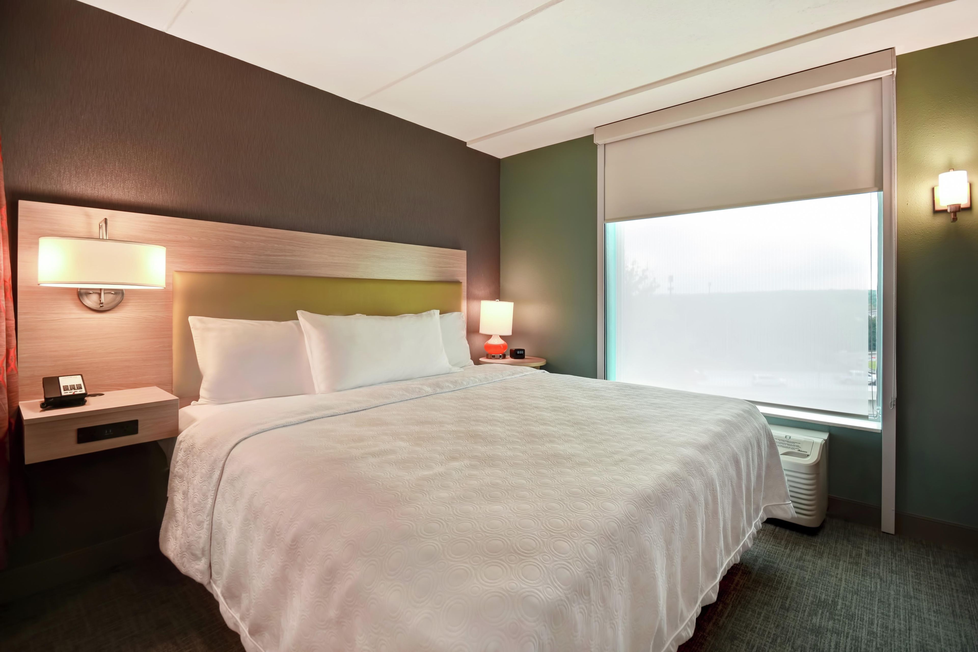 Una habitación de hotel con una cama, mesitas de noche, lámpara y una ventana con persianas.