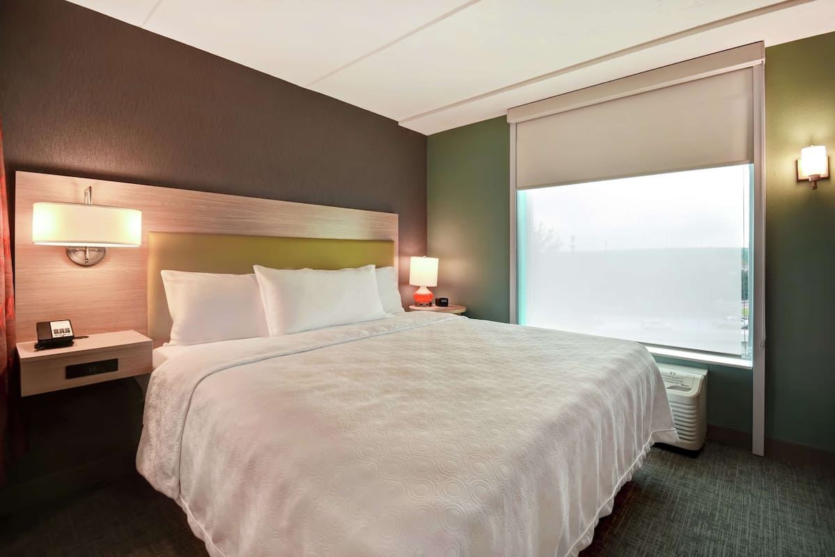 Une chambre d’hôtel avec un grand lit, une table de chevet avec une lampe, une fenêtre avec des stores et un luminaire fixé au mur.