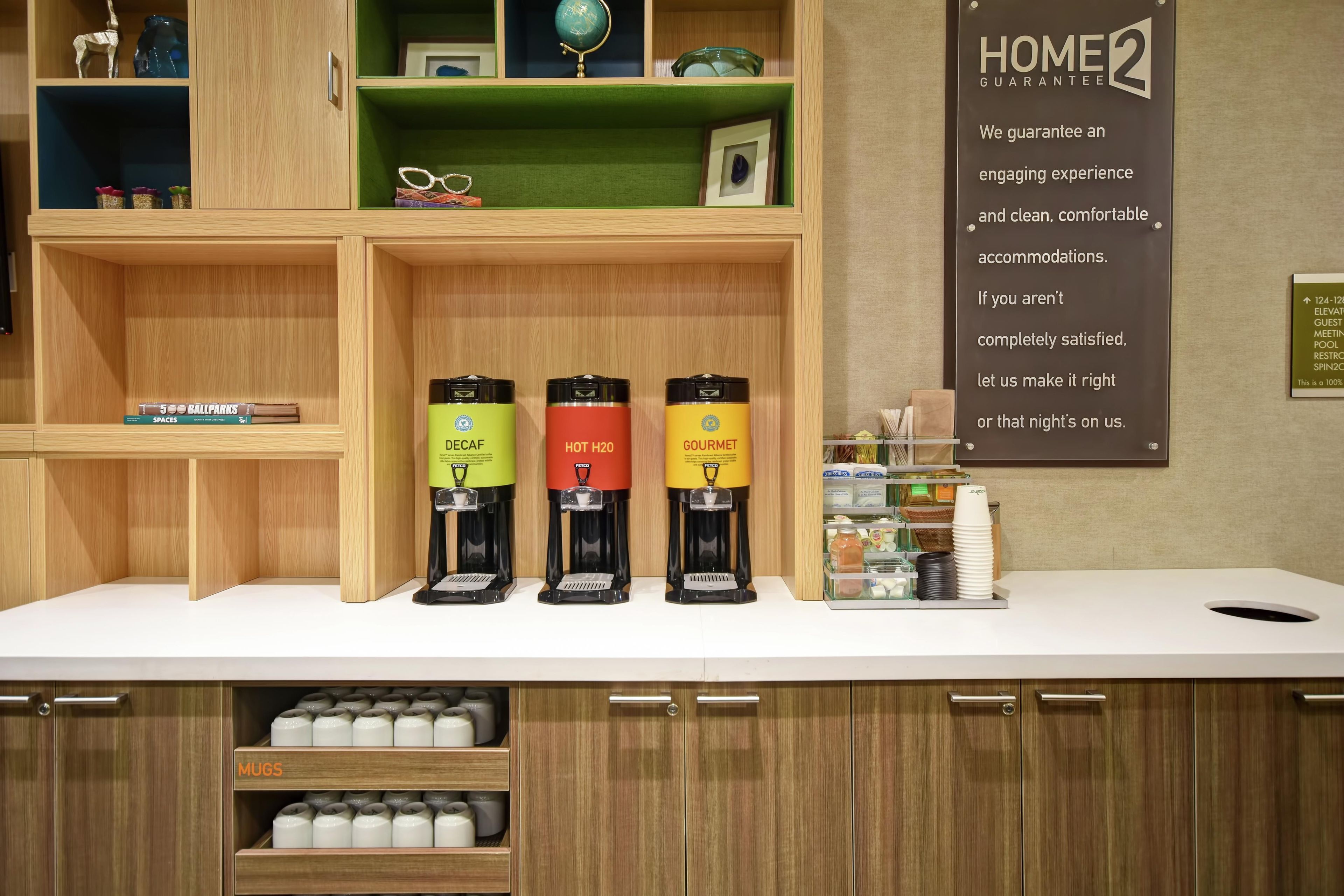 A hotel reception area with a counter displaying three beverage dispensers labeled 'DECAF,' 'HOT H2O,' and 'GOURMET.' There are shelves above with various items, and a sign on the wall to the right.