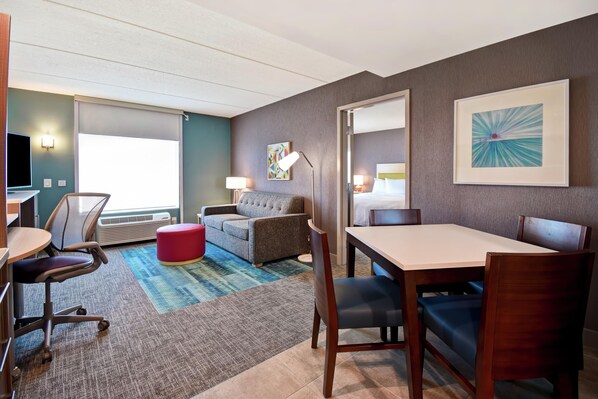 Habitación de hotel con sofá, mesa de comedor, sillas, televisor, lámpara y un cuadro en la pared.