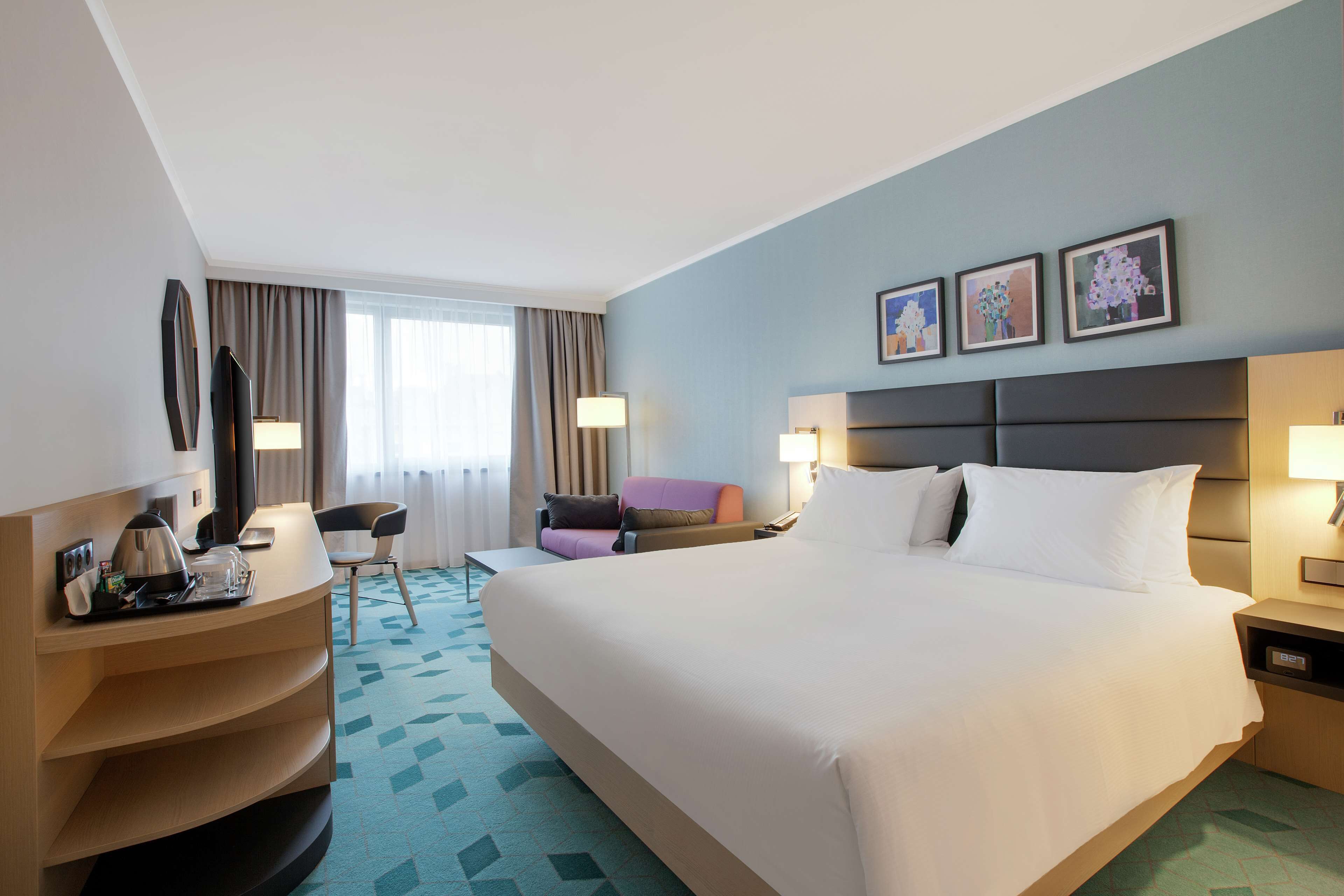A hotel room with a large bed, a desk with a coffee maker, a chair, and a window with curtains.
