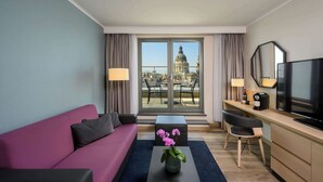 Habitació d'hotel moderna amb una finestra gran que ofereix vista a un edifici històric, un sofà confortable, una taula petita amb un ram de flors, un escriptori amb televisió i una cadira.