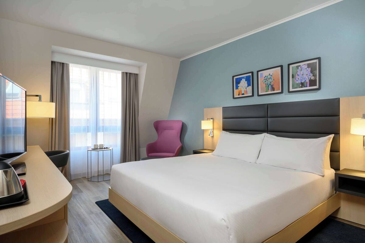 Una habitación de hotel con una cama grande, un escritorio con una computadora portátil, una silla rosa y obras de arte en la pared.