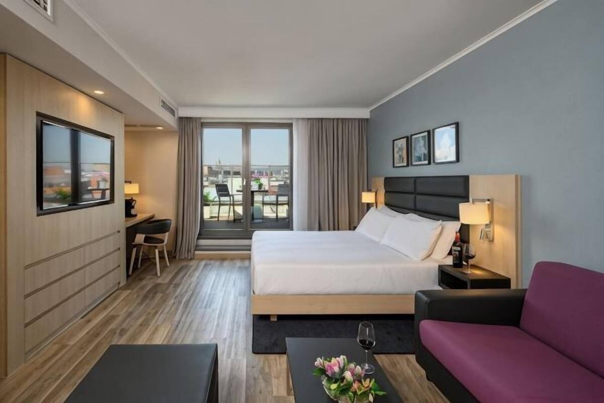 Habitación de hotel con cama, escritorio, silla, sofá, televisor y balcón con vistas.