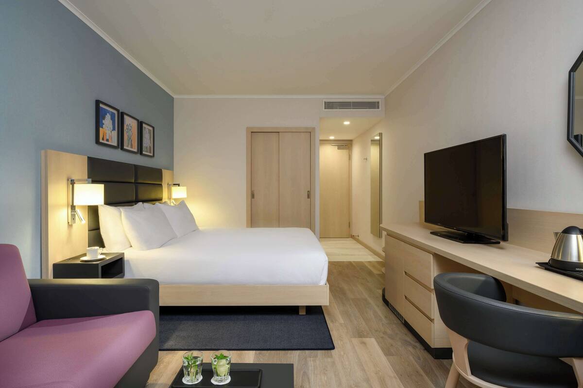 Habitación de hotel con cama, mesita de noche con lámpara, escritorio con televisor, una silla y una pequeña mesa con vasos.