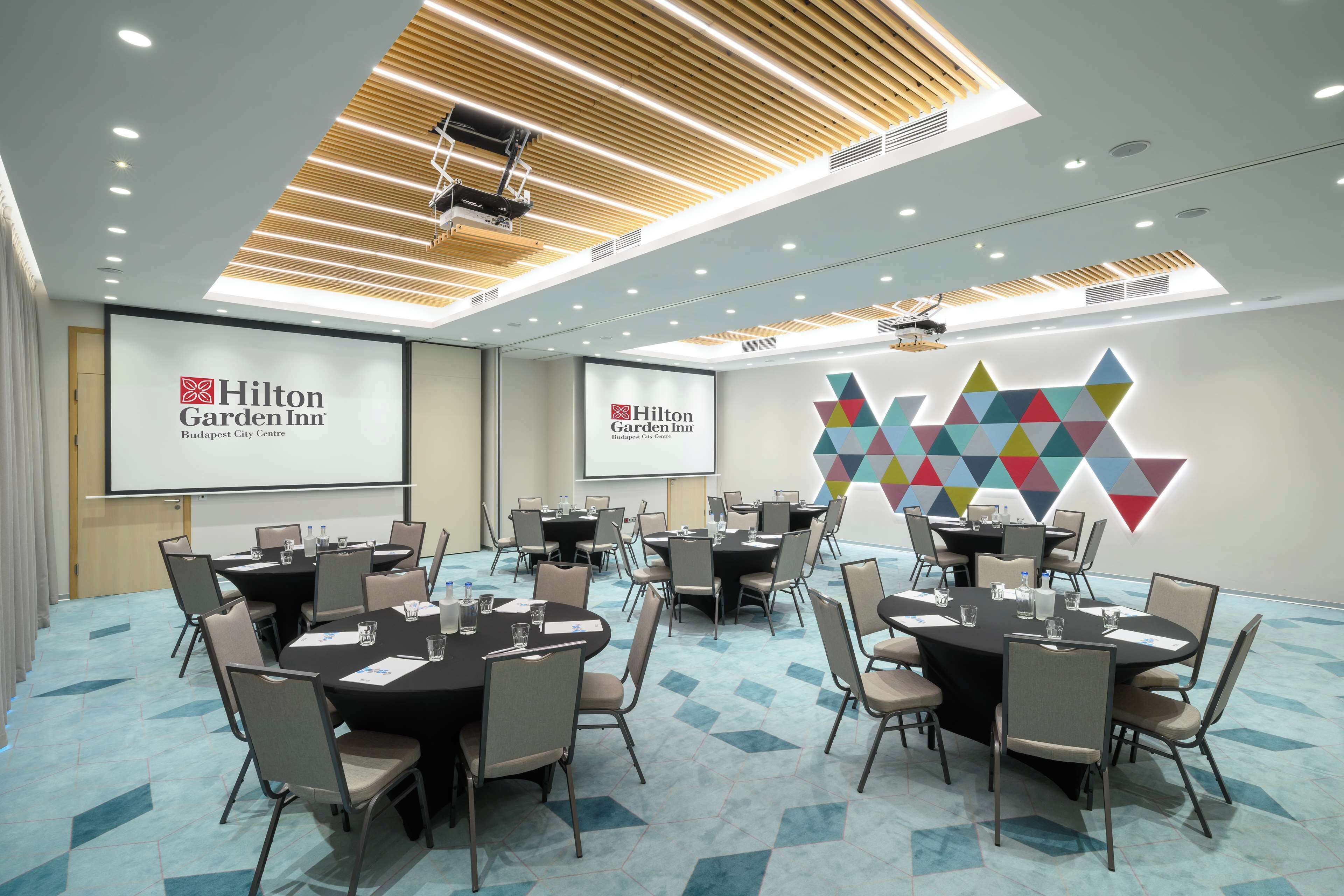 A conference room at Hilton Garden Inn with multiple tables and chairs, a projector screen, and a geometric wall decoration.