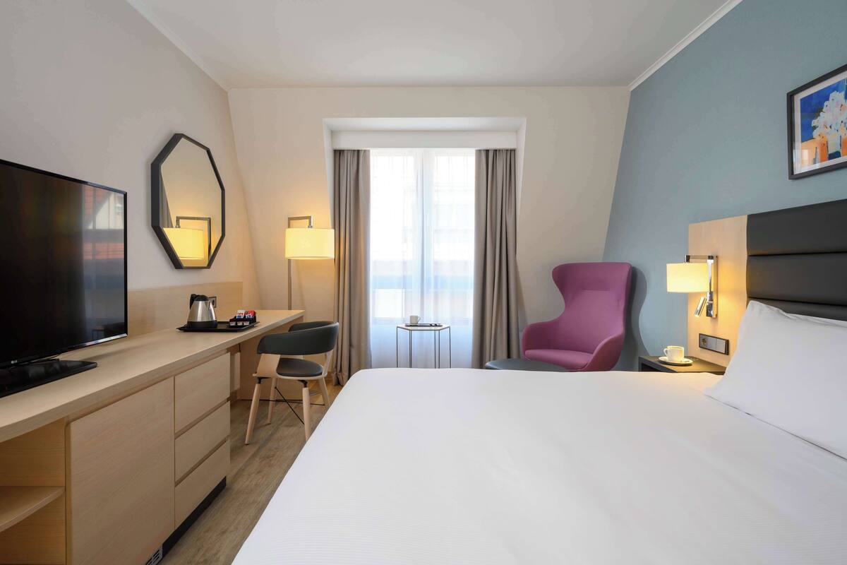 Habitación de hotel con cama, escritorio con televisor, silla, mesita pequeña y ventana con cortinas.