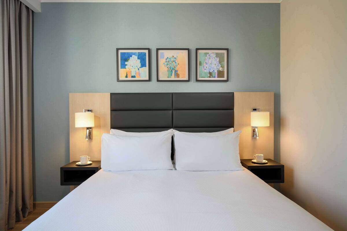 Una habitación de hotel con una cama grande, dos mesitas de noche con lámparas, y tres cuadros enmarcados en la pared.