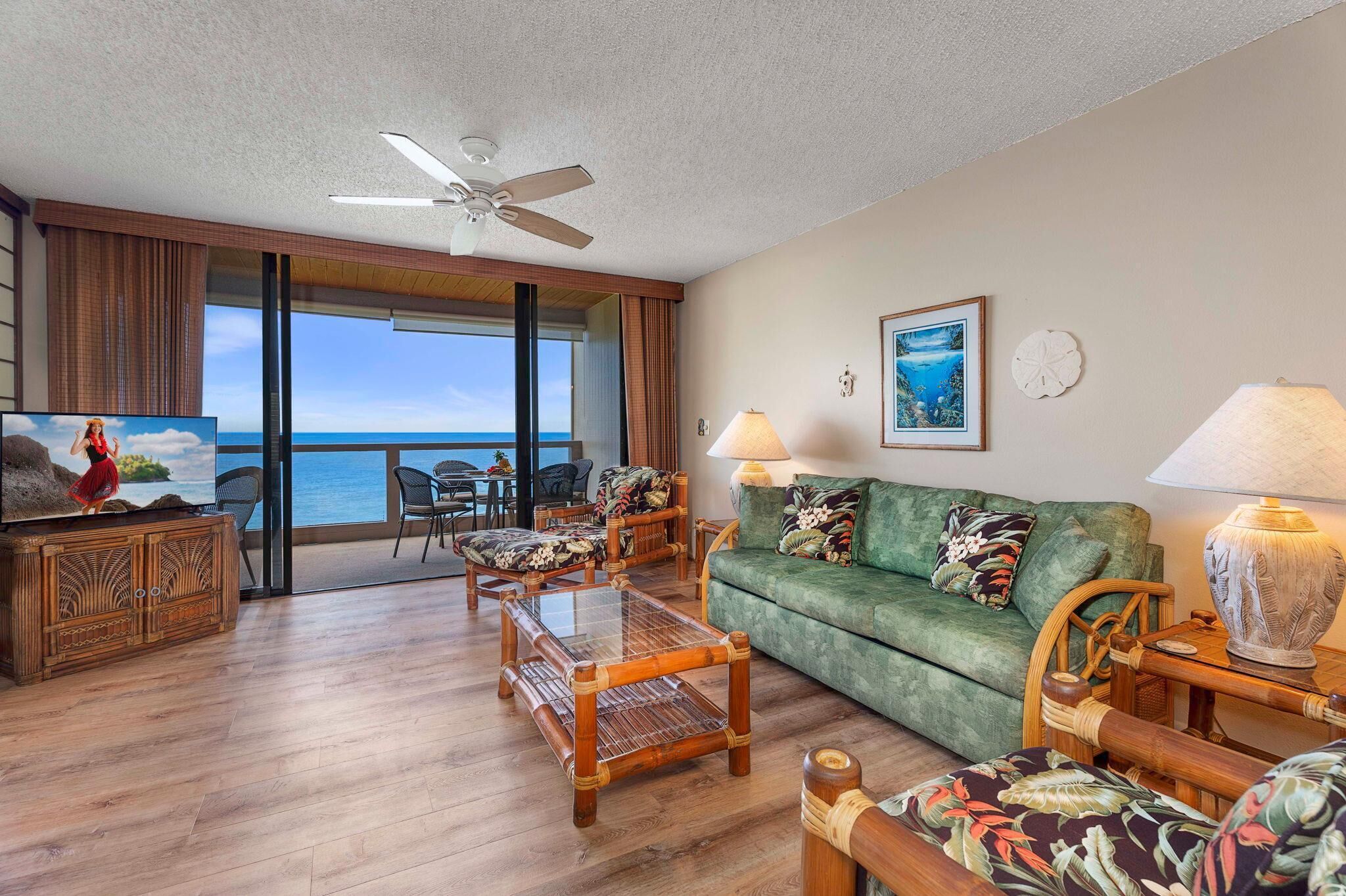 A spacious living room with a green sofa, wooden coffee table, and chairs. There is a television on a wooden cabinet, a ceiling fan, and a large window with a view of the ocean.