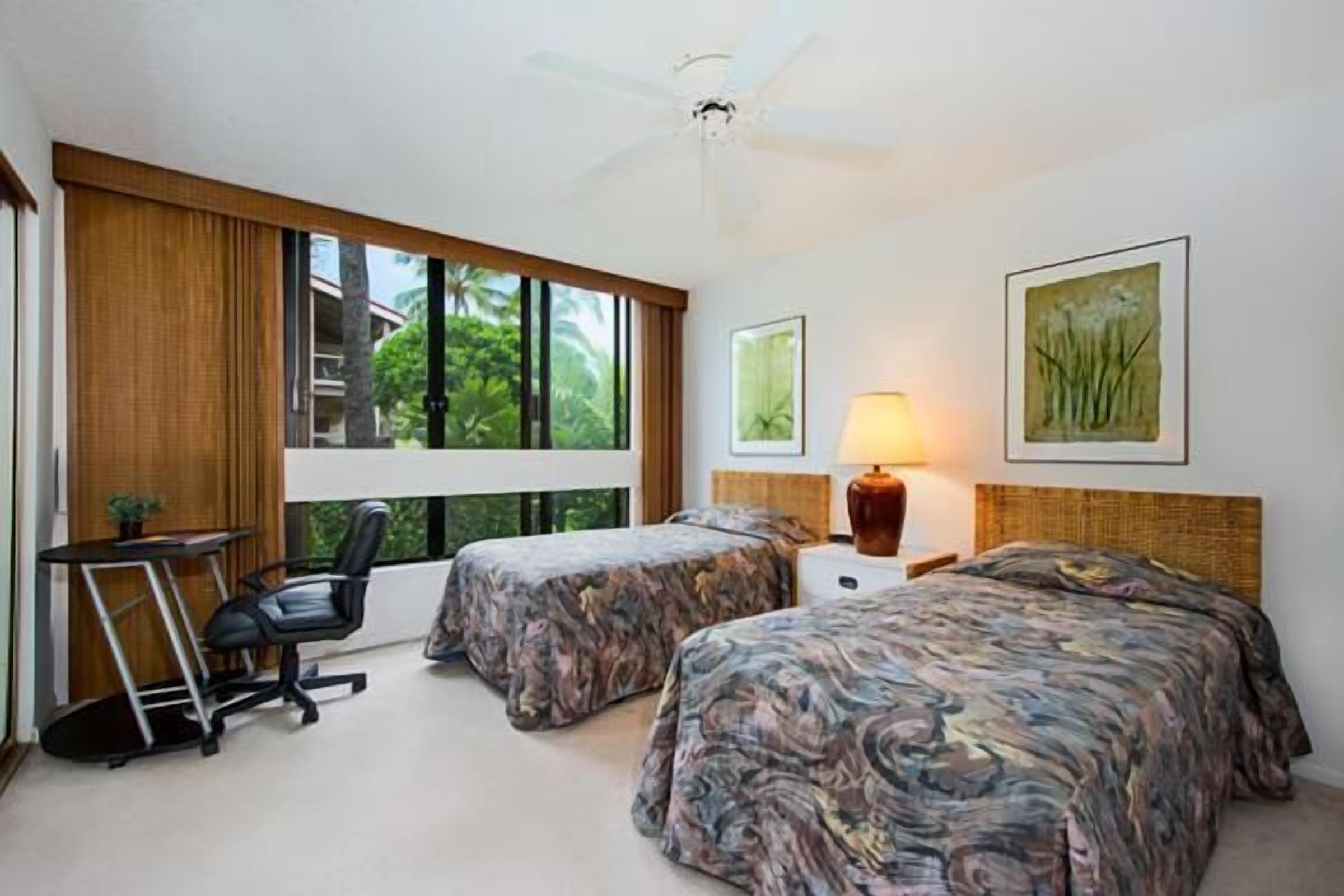 A hotel room with two beds, a desk, a chair, and a window with a view of greenery.