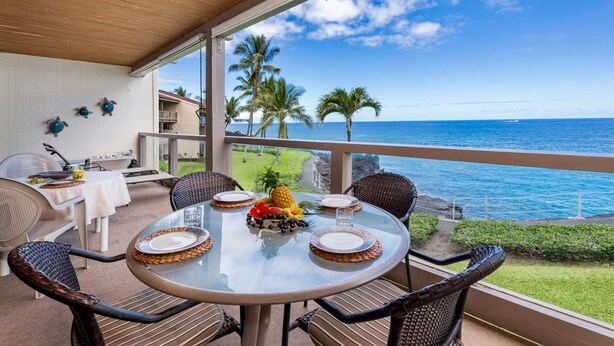 A round glass table with white tablecloths and woven placemats, surrounded by wicker chairs, overlooking a beach with palm trees and the ocean. - Keauhou Kona Surf & Racquet Club 3202 (Kailua-Kona)