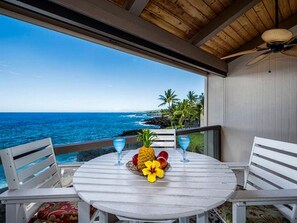 A round table with a fruit bowl and two glasses, overlooking a coastal view with palm trees and the ocean. - Keauhou Kona Surf & Racquet Club #5-303 (Kailua-Kona)