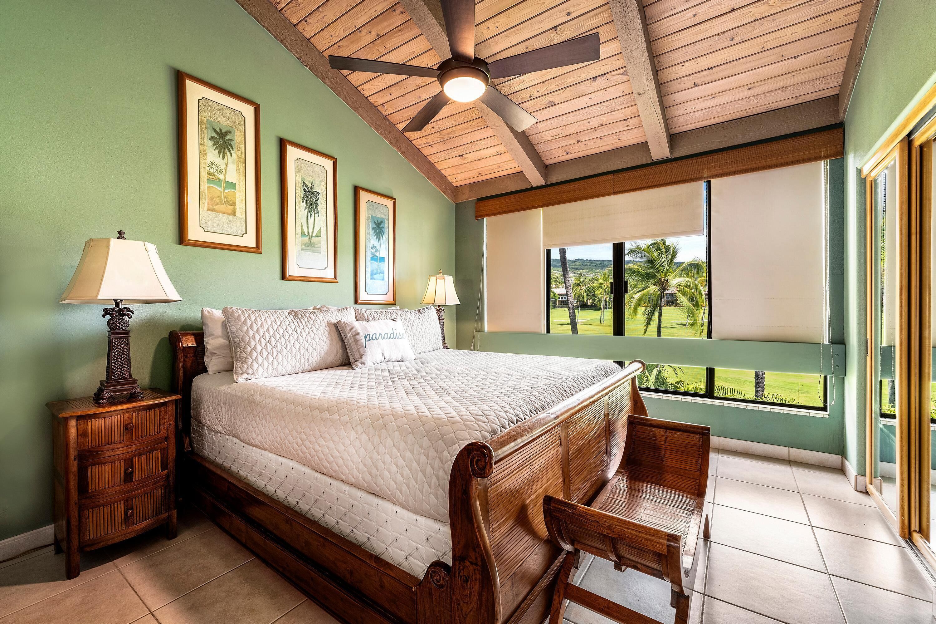 Une chambre à coucher avec un grand lit, une tête de lit en bois, deux tables de chevet avec des lampes, des tableaux encadrés au mur et une vue sur l’extérieur par une fenêtre.