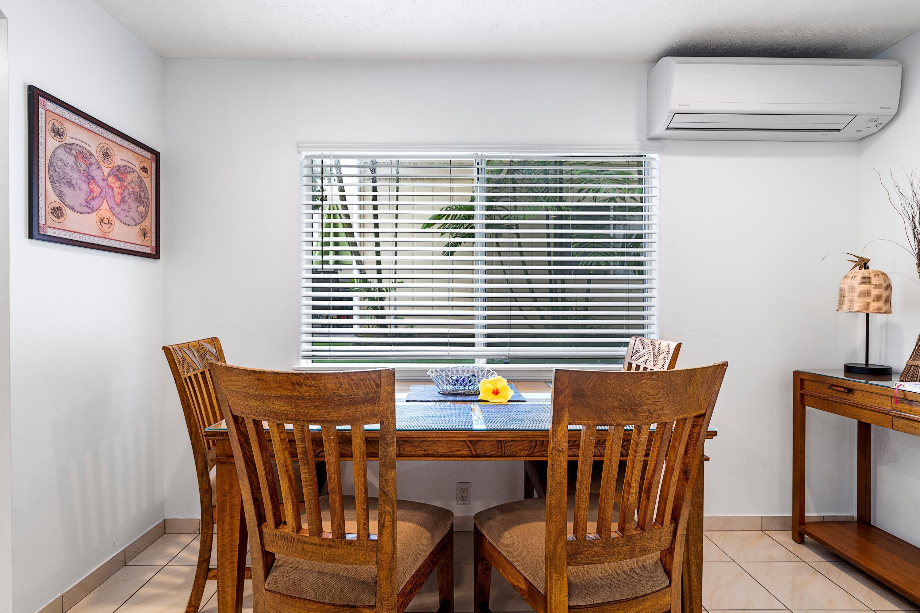 A dining area with a wooden table and chairs, a window with blinds, a wall-mounted air conditioner, and a framed picture.