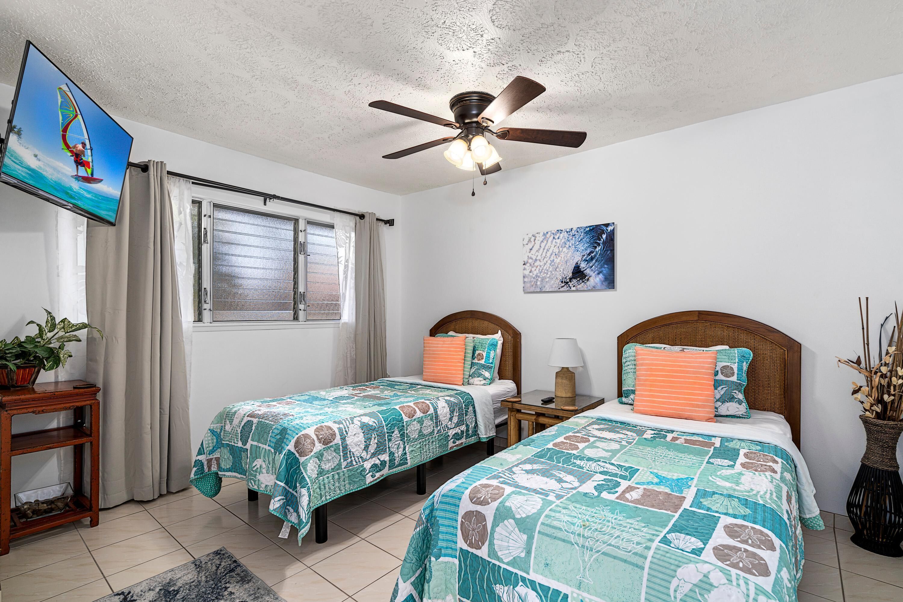 A hotel room with two beds, a ceiling fan, a TV, a window with curtains, a painting, and a small table with a lamp.