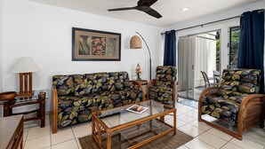 Un salon comprenant un canapé, des fauteuils, une table basse, un ventilateur de plafond, un tableau encadré et une vue sur l’extérieur, visible à travers une porte vitrée.