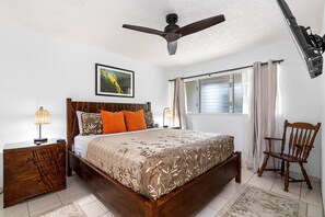 Un dormitorio con cama de madera, ventilador de techo, mesita de noche con lámpara, una silla y ventana con cortinas.