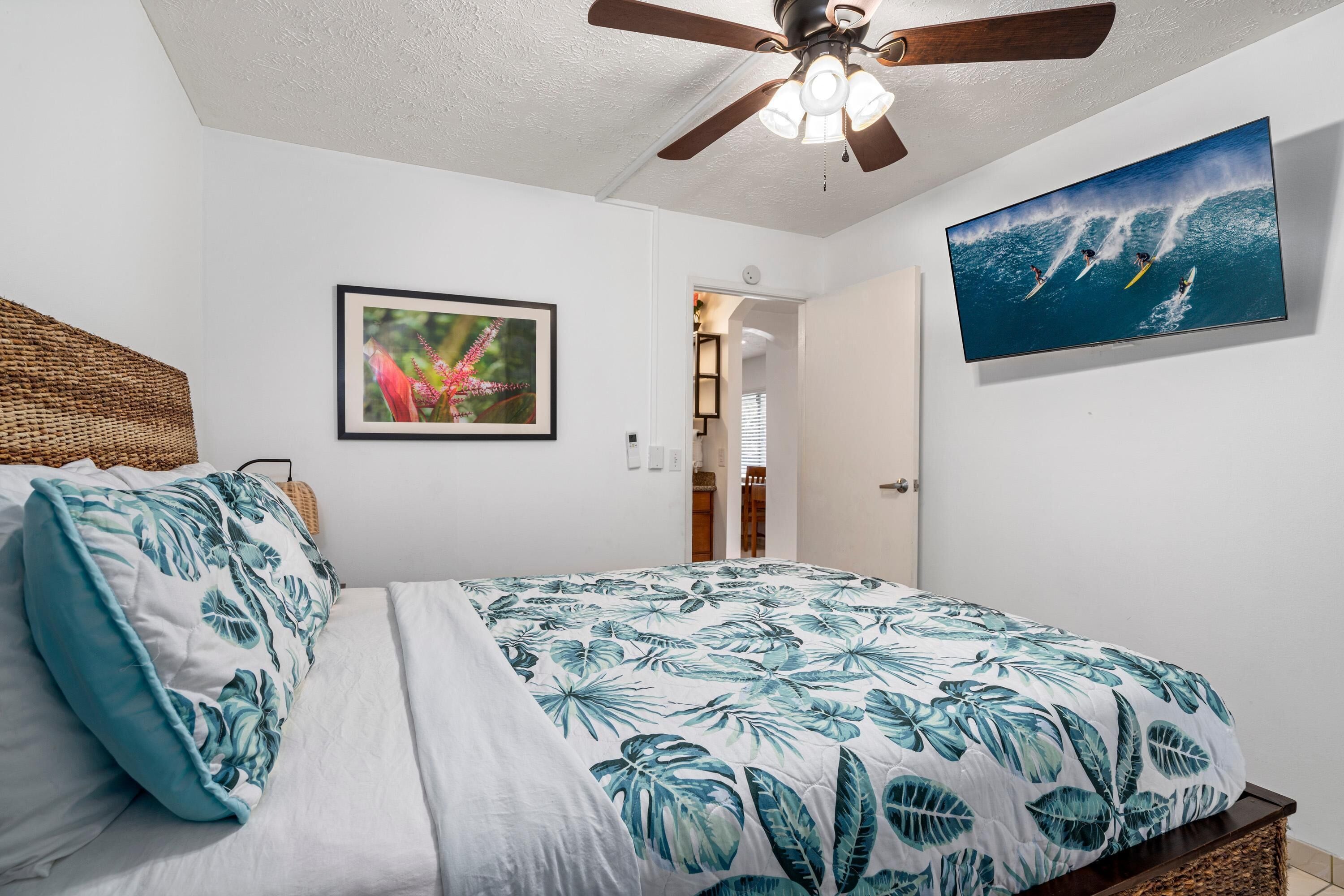 Ein Schlafzimmer mit einem Bett, einem Deckenventilator, einem gerahmten Bild und einem Fernseher, der eine Surfszene zeigt.