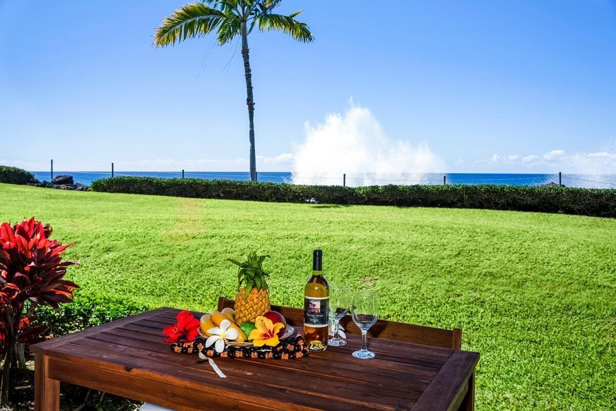 Una mesa de madera con una botella de vino, una copa y frutas tropicales sobre un césped con una palmera y vista al océano.