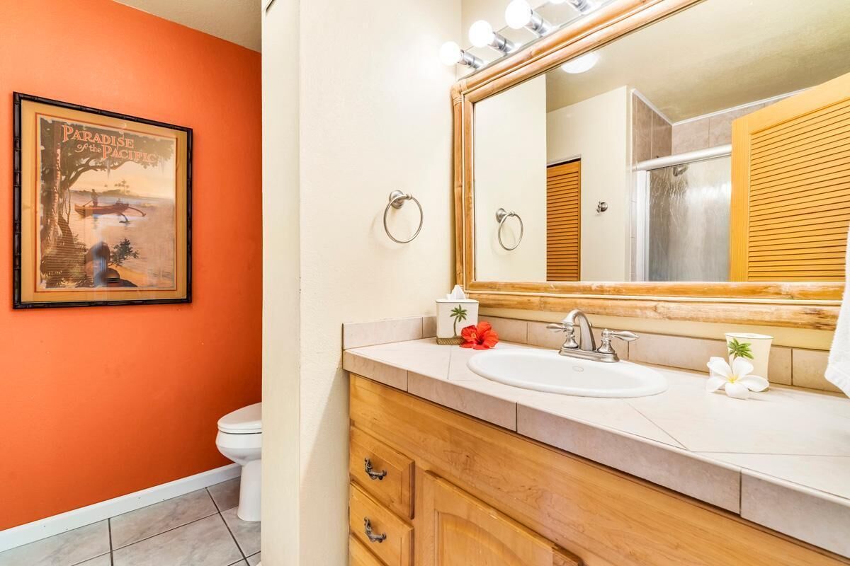 A bathroom with an orange wall, a wooden vanity, a large mirror with lights, a sink, and a toilet.
