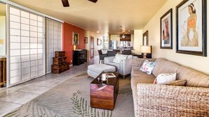 Ein geräumiges Wohnzimmer mit einer Couch, Sesseln, einem Couchtisch und einem Essbereich mit Tisch und Stühlen. Es gibt einen Deckenventilator, eine Glastür mit Schiebeöffnung sowie gerahmte Kunstwerke an den Wänden.