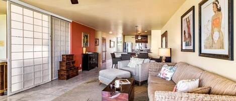 Amplia sala de estar con sofá, sillones, una mesa de centro y un comedor con mesa y sillas. Hay un ventilador de techo, una puerta corrediza de vidrio y cuadros en las paredes.