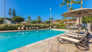 Ein Swimmingpool mit Liegestühlen und Sonnenschirmen, umgeben von Palmen und blauem Himmel.