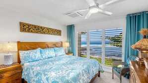 A bedroom with a bed, a ceiling fan, a nightstand, a chair, and a view of the ocean through large windows.